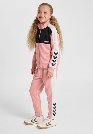 Pinker Trainingsanzug mit schwarzen Akzenten, bestehend aus einer Reißverschlussjacke mit Mesh-Stoff und weißen Streifen, kombiniert mit konisch zulaufenden Hosen und sportlichen Schuhen.