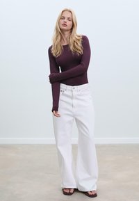 Langärmeliges, burgunderfarbenes, figurbetontes Oberteil, kombiniert mit hoch taillierten weißen, weiten Jeans und braunen flachen Sandalen vor einem neutralen Hintergrund.