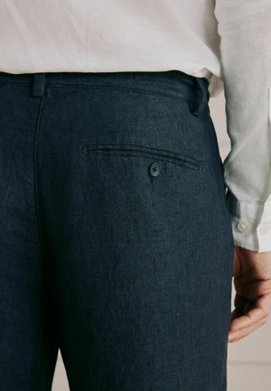 Mann trägt dunkelblaue Leinenhose mit einer geknöpften Gesäßtasche und einem weißen langärmeligen Hemd, das in die Hose gesteckt ist.