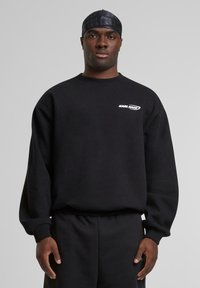 Black sweatshirt with a crew neck, featuring a white "KARL KANI" logo on the chest. Fabric appears soft and textured. Long sleeves with ribbed cuffs.