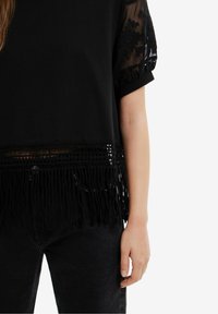 Černý top s krátkými rukávy vyrobený z průhledné vyšívané látky, s dekorativními třásněmi na spodním okraji a složitými detaily podél okrajů.