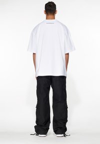 T-shirt bianco di cotone oversize con maniche corte, caratterizzato da un logo discreto sulla parte posteriore. Abbinato a pantaloni cargo neri con tasche.