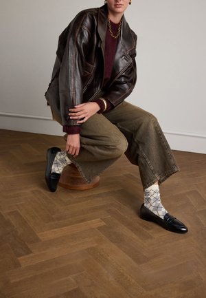 Bruine leren jas over een kastanjebruine trui, olijfgroene wijde broek, zwarte loafers en geometrisch patroonsokken op een houten vloer.