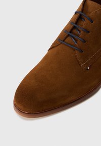 Chaussure habillée en daim marron avec des lacets bleu foncé, des coutures visibles et une petite étiquette en tissu près des oeillets sur un fond blanc.