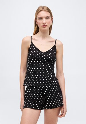 Vrouw draagt een zwarte mouwloze top met polka dots en bijpassende korte broek, staand tegen een effen lichte achtergrond.