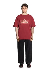 Czerwona bawełniana koszulka z logo "Prosto" w kolorze złotym, luźny krój, zestawiona z luźnymi czarnymi dżinsami i czarnymi trampkami z czerwonymi akcentami.
