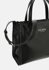 Schwarze Handtasche aus Synthetik mit doppelten Griffen, silbernem Logo "GUESS EST 1981" und kontrastierenden cremefarbenen Nähten entlang der Kanten.