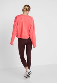 Korálový svetr s kleslými rameny, doplněný tmavě burgundskými legínami. Modelka má na sobě bílé sportovní boty. Jednoduchý design látky, hladká textura.