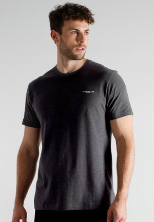 Man with curly hair looking right, wearing a fitted dark gray T-shirt with small "Tom Tailor" logo on chest against plain background.