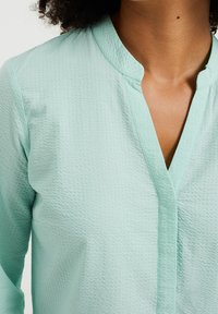 Světle mátově zelená košile s texturovaným povrchem, vybavená mandarinovým límcem a předními klopy, které dodávají jemný detail.