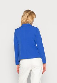 Blazer structuré bleu avec col cranté, manches longues, coupe ajustée et texture lisse, assorti à un pantalon blanc.