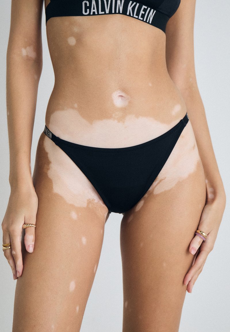 Črni bikini Calvin Klein z minimalnim dizajnom, gladko teksturo in kontrastom s kožo z izrazitimi svetlejšimi lisami.