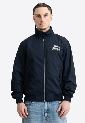 Mies, jolla on letitetyt hiukset ja päällään vetoketjullinen laivaston sininen takki, jossa on Lonsdale Londonin logo, sekä vaaleansiniset farkut yksivärisen taustan edessä.