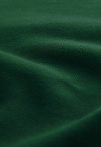 Donkergroene katoenstof met een gladde textuur, die subtiele plooien en een uniforme weefpatroon toont. Zacht en licht van uitstraling.
