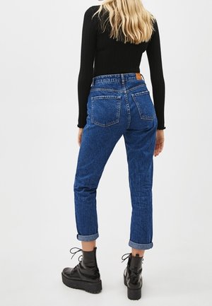 Blondynka w podwiniętych niebieskich dżinsach, czarnej prążkowanej bluzce z długim rękawem i czarnych platformowych botkach na sznurowadła, stojąca tyłem.