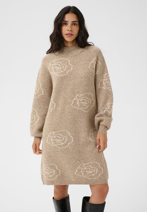 Donna dai lunghi capelli scuri che indossa un vestito beige a maglia con motivi floreali bianchi e stivali neri al ginocchio, in piedi davanti a uno sfondo semplice.