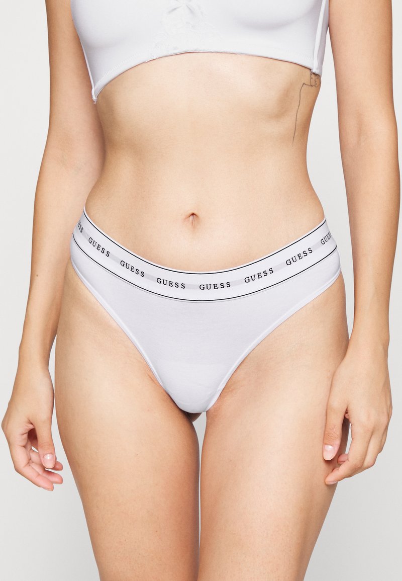 String en coton blanc avec une ceinture logo "GUESS" en noir et blanc, présentant une texture lisse et un design à couverture minimale.