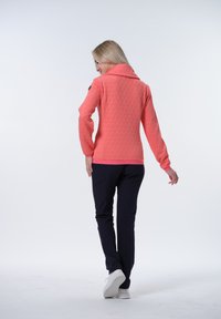 Koraalkleurige gewatteerde jas met een hoge kraag, voorzien van een geometrisch patroon en een roze zoom, gecombineerd met marineblauwe broek en witte sneakers.