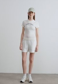 Λευκό βαμβακερό t-shirt με το λογότυπο «TOMMY JEANS», συνδυασμένο με λευκά σορτς. Το μοντέλο φοράει ανοιχτό πράσινο καπέλο και λευκά αθλητικά παπούτσια.