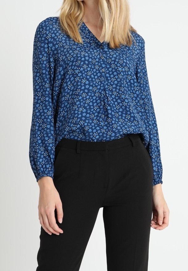 Femme portant un chemisier bleu à manches longues avec de petites fleurs blanches et un pantalon noir sur mesure, debout devant un fond uni.