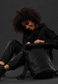 Schwarzer Rolltop-Rucksack aus strukturiertem Material, mit seitlichem Reißverschluss, verstellbaren Riemen und einem geprägten Logo. Minimalistisches Design.