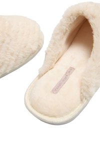 Zapatillas beige difusas con interior y exterior suaves. Forma redondeada, bordes elásticos y suela de color claro. Etiqueta visible en el interior.