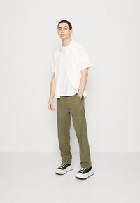 Kortärmad vit skjorta över en vit T-shirt, ihop med olivgröna byxor och svart-vita sneakers, stående mot en neutral bakgrund.