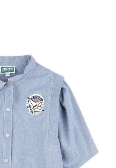 Chemise en jean bleu à manches courtes avec un patch rond représentant une grue en origami blanche et un texte, fermeture à boutons et texture douce.