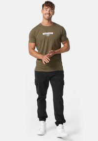 Olivgrünes T-Shirt mit weißem, aufgedrucktem Logo, kombiniert mit schwarzen Cargohosen mit Seitentaschen und weißen Sneakers.