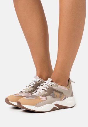 Jambes portant des baskets épaisses à motif camouflage beige, marron et rose avec des lacets blancs et des semelles blanches épaisses sur fond blanc.