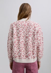 Frau, die von hinten schaut, trägt einen pink-weißen Leopardenmuster-Pullover und pinke Zebra-Muster-Hosen vor einem einfachen hellen Hintergrund.