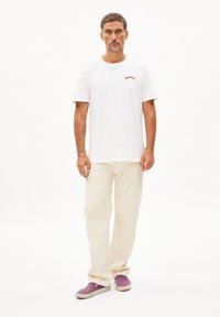 Witte katoenen T-shirt met rode geborduurde tekst, beige wijde broek en paarse canvas sneakers met witte zolen en grijze accenten.