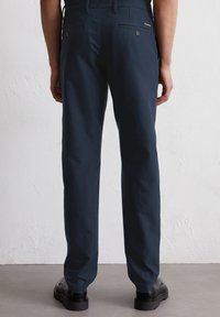 Navyblauwe broek met een rechte snit, voorzien van twee achterzakken met knopen en een gestructureerde afwerking. Zwarte, platte schoenen worden gedragen.