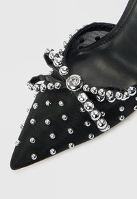 Melns, ar teksturētu audumu izgatavots, ar smailu purngala daļu kurpes, rotātas ar sudraba kniedēm un dekoratīvu loku priekšpusē. Gluda iekšējā oderējuma kārta.