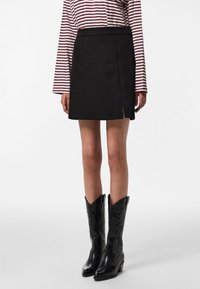 Femme portant une mini-jupe noire avec une fente à l'avant, un haut rayé à manches longues et des bottes noires jusqu'aux genoux de style western, debout sur fond blanc.