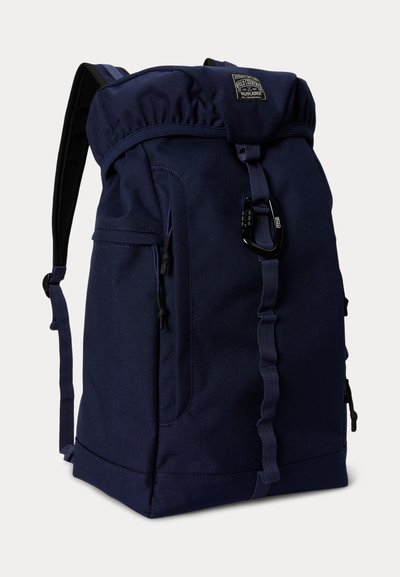 Σακίδιο πλάτης από ύφασμα σε σκούρο μπλε χρώμα με εμπρόσθια τσέπη με φερμουάρ, ρυθμιζόμενους ιμάντες, επάνω κάλυμμα που κλείνει με καραμπίνερ και μικρό διακοσμητικό με το λογότυπο της μάρκας.