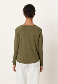 Kõrvas lokkis juustega naine, kellel on kuklis soeng, seljas oliivsest rohelisest pika varrukaga särk ja valged teksad, seisab tavalise heleda tausta ees.