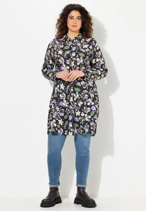 Knielange, met knopen sluitende jurk met lange mouwen in zwarte stof versierd met witte, paarse en groene bloemenpatronen; gecombineerd met blauwe skinny jeans en zwarte laarzen.