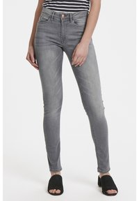 Jeans ajustados grises hechos de denim con un acabado desgastado, que cuentan con cinco bolsillos, pasadores de cinturón y un cierre de botón. Con zapatos negros de punta abierta.