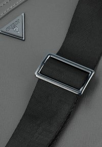 Grau gemustertes Material mit einem silbernen Logo-Dreieck und einem schwarzen Riemen mit einer verchromten rechteckigen Schnalle. Strukturierte Oberflächendetails sind sichtbar.