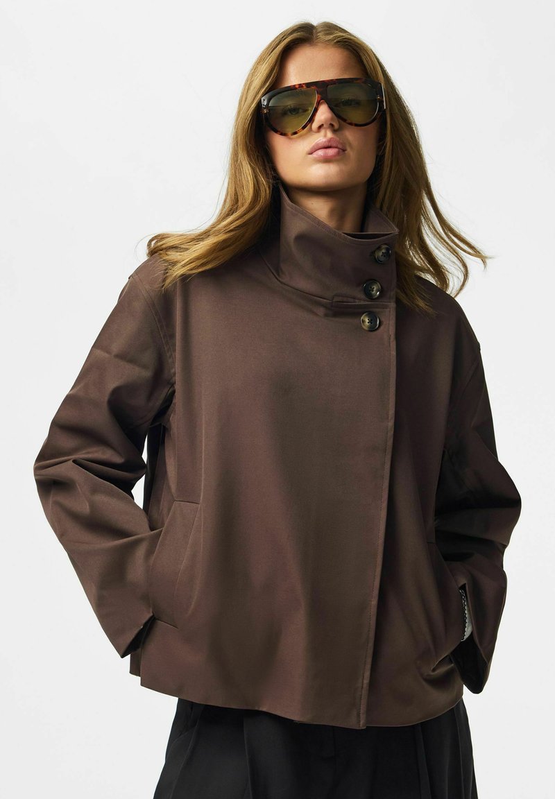 Femme portant de grandes lunettes de soleil écaille de tortue et une veste marron à col montant avec des boutons, mains dans les poches, sur fond uni.