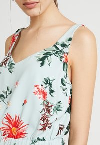 Blommig linnetopp med ljusblå bakgrund, röda och gröna blommönster, och en v-ringning med breda axelband.