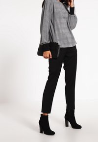 Donna che indossa una blusa grigia a quadri con maniche lunghe, pantaloni neri a sigaretta, stivaletti neri con tacco, e tiene in mano una borsa a pochette nera.