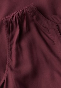 Burgundské tričko vyrobené z jemného a hladkého materiálu, které má nařasený výstřih a čisté švy po okrajích. Bez viditelných vzorů nebo kovových doplňků.