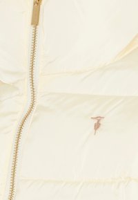 Crème pufferjack met gouden rits, voorzien van een gequiltde textuur en een klein geborduurd logo aan de linkerkant.