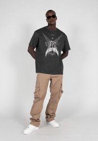 Mørkegrå grafisk t-shirt med vingertryk og tekst, parret med beige cargo-bukser med flere lommer og hvide sneakers.