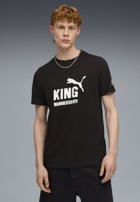 Camiseta de algodón negra con texto blanco "KING" y el logo de "MANCHESTER CITY", con un logo blanco de Puma en el lado derecho y mangas cortas.