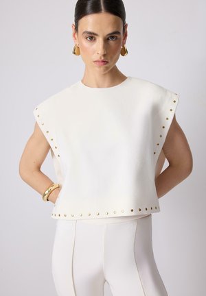 Žena s účesem uhlazeným dozadu, která má na sobě bílý top bez rukávů se zlatými cvoky, bílé přiléhavé kalhoty, zlaté náušnice a zlatý náramek.