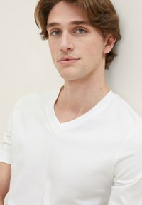 Weißes Baumwoll-T-Shirt mit V-Ausschnitt, kurzen Ärmeln und glatter Textur, getragen von einem Model vor einem hellen Hintergrund.
