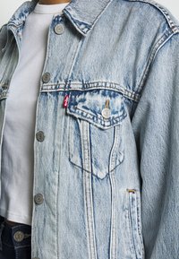 Ljusblå denimjacka med silverfärgad knappar, två framfickor och en röd etikett. Har en klassisk krage och synliga stygn.